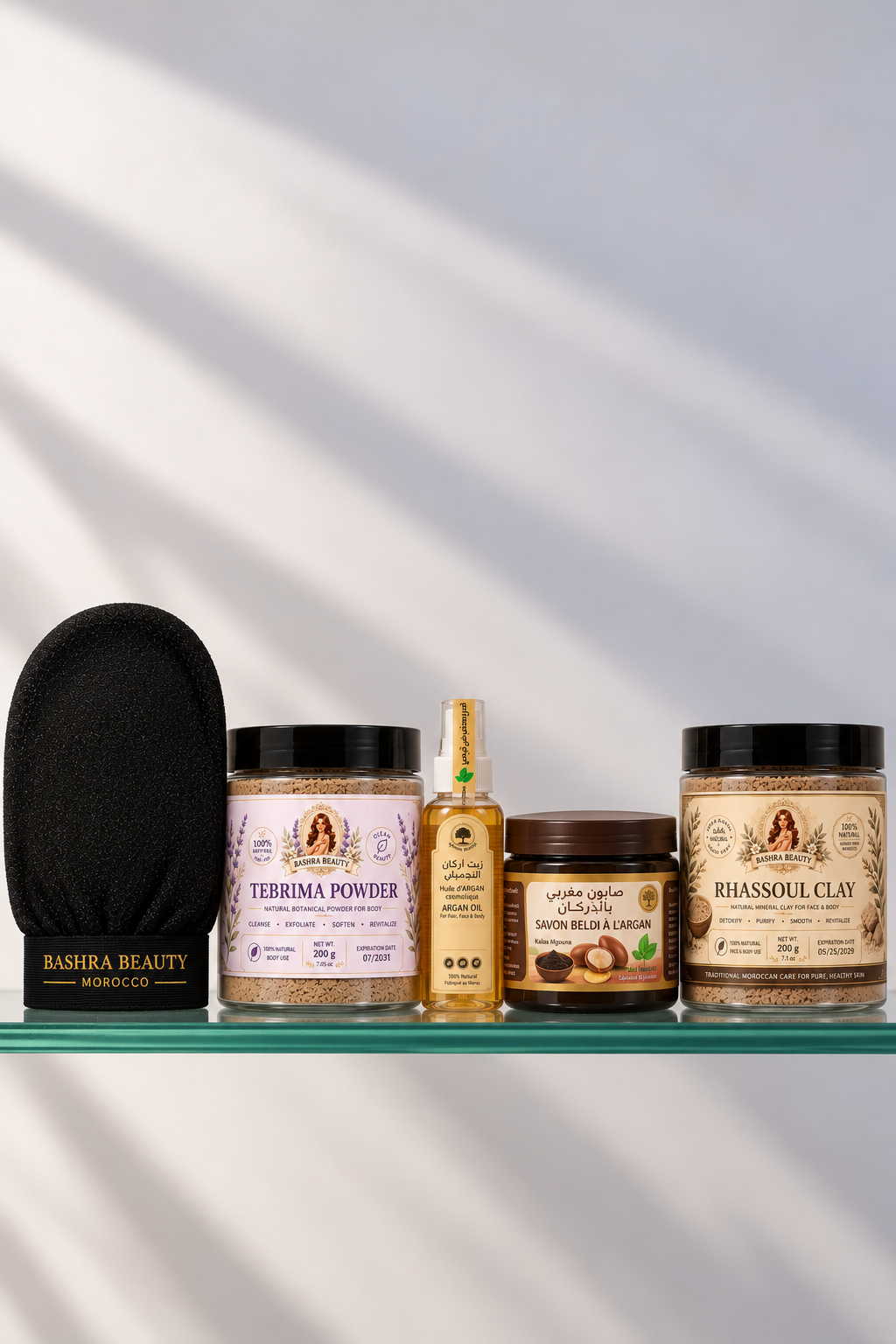 Bashra Beauty Moroccan body care products arranged on a glass shelf, featuring Tebrima Powder, argan oil, Moroccan black soap, rhassoul clay, and a traditional exfoliating glove.