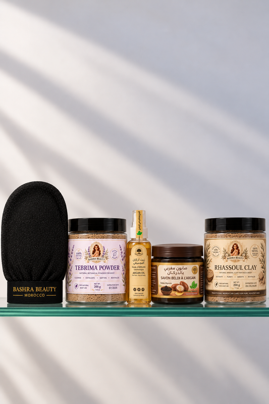 Bashra Beauty Moroccan body care products arranged on a glass shelf, featuring Tebrima Powder, argan oil, Moroccan black soap, rhassoul clay, and a traditional exfoliating glove.